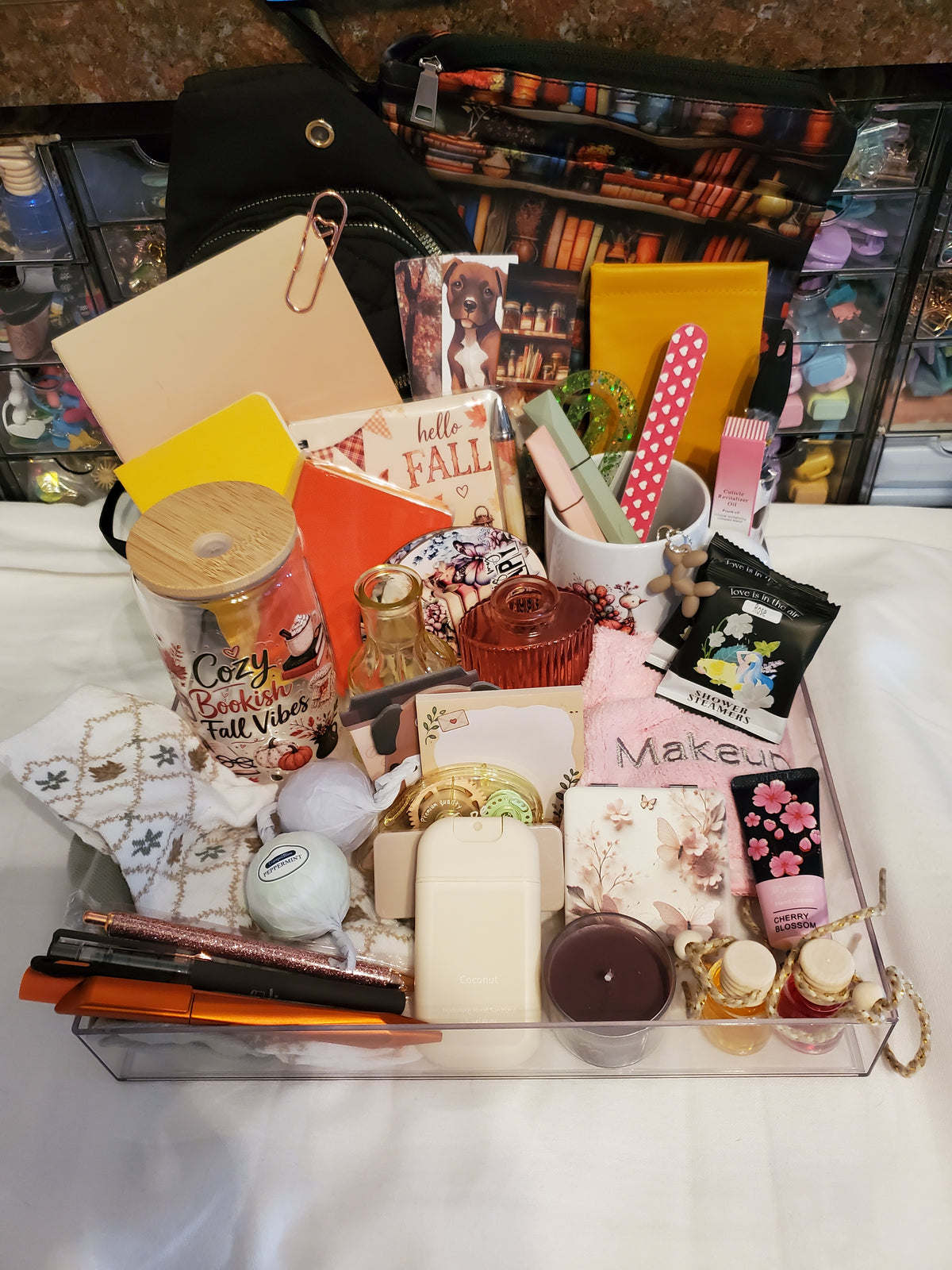 A clear acrylic organizer tray filled with assorted fall-themed and beauty items, including a mug with pink polka-dot straws, a candle, lip gloss, a notebook, and other stationery and personal care accessories, set on a white surface with a black bag and shelves in the background.