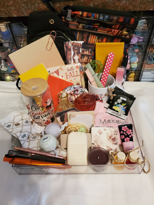A clear acrylic organizer tray filled with assorted fall-themed and beauty items, including a mug with pink polka-dot straws, a candle, lip gloss, a notebook, and other stationery and personal care accessories, set on a white surface with a black bag and shelves in the background.